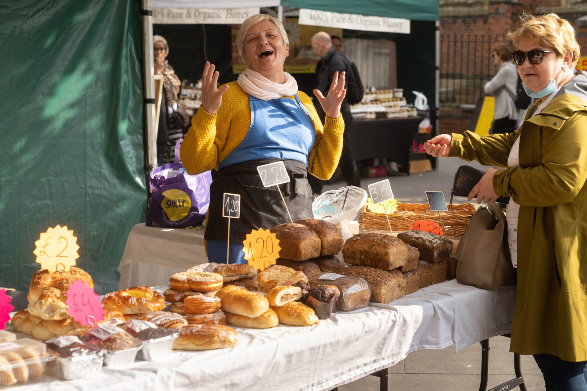 A woman wearing a mustard-coloured jumper and blue and black apron stand behind a table fully of freshly baked bread and cakes. She's laughing with her hands raised.