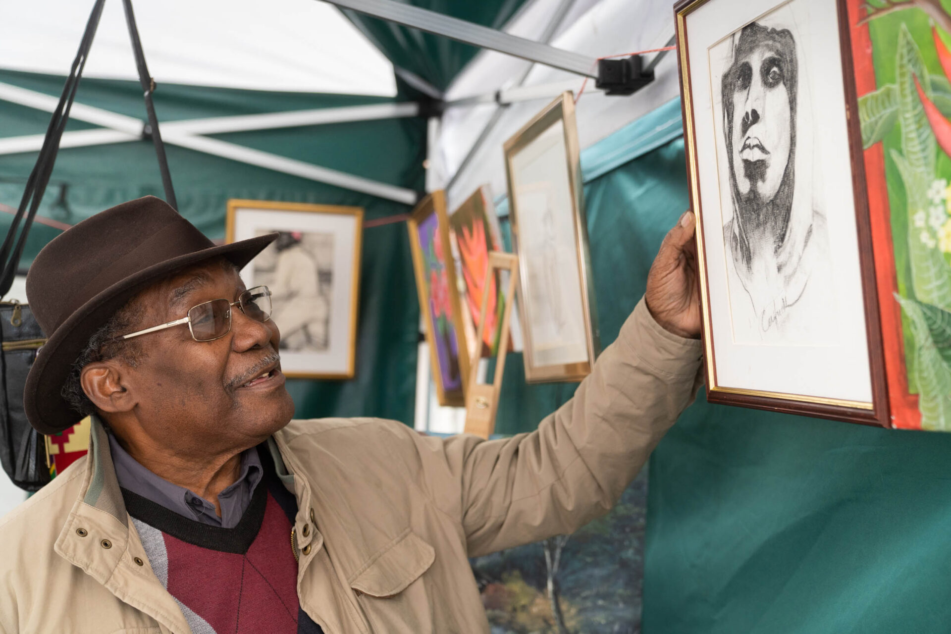 An older black man wearing a brown trilby hat, beige jacket, dark red v-neck jumper and grey shirt, holds the edge of a framed picture at an art stall.