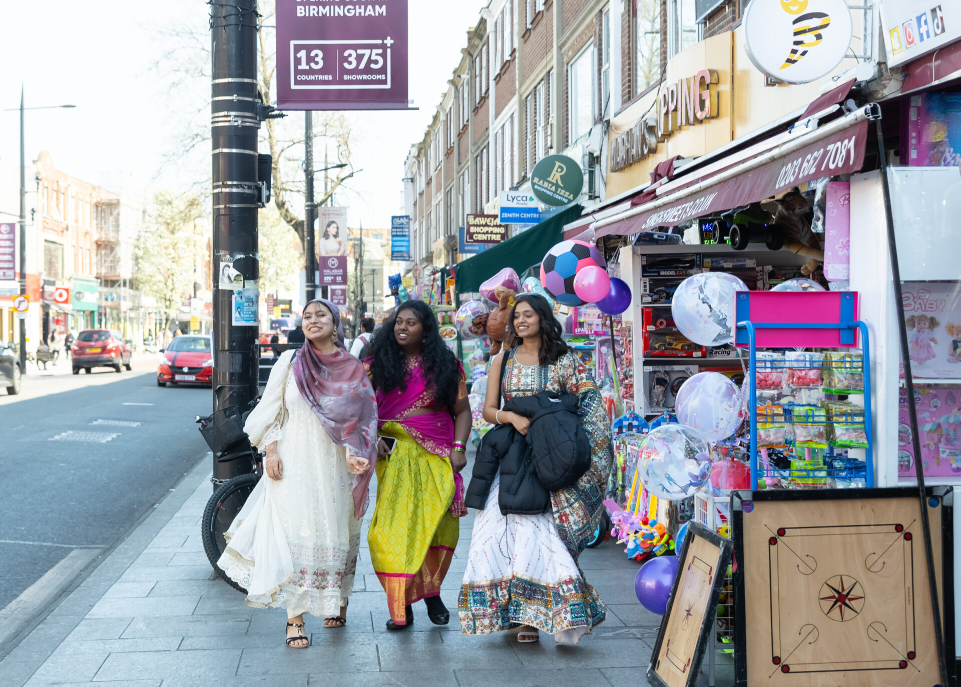 3 young women walk down Southall Highstreet wearing brightly coloured Saris.