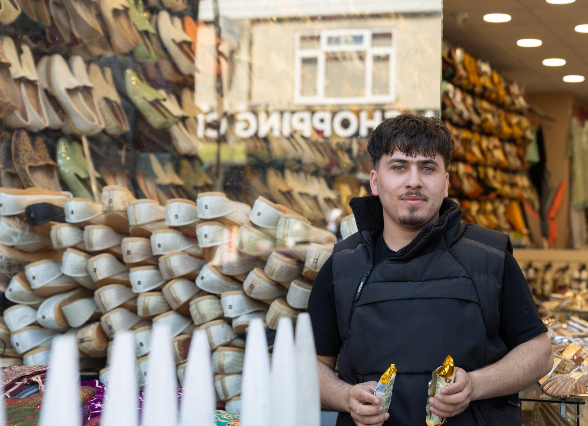 A young man with dark brown hair, wearing a black t-shirt holds 2 ice creams while standing in front of a shop window.
